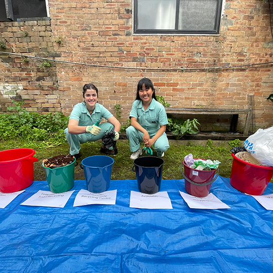Two people crouching behind buckets sorted and labelled with different types of waste materials