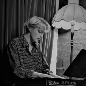 Black and white image of a solo pianist playing at a keyboard next to an older styled lamp with moody lighting