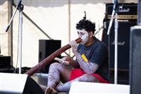 A young First Nations person sits on stage wearing a black shirt, red shorts and ochre whilst playing the yidaki (didgeridoo)