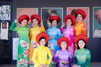 A group of eight women dressed in brightly coloured gowns and wearing traditional Vietnamese red head pieces pose for a photo against a wall full of paste ups