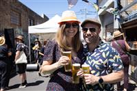 Two happy people wearing hats and sunglasses, holding cold beverages standing in a sunny laneway with other people seen in the background