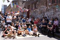 A packed sunny laneway decorated with colourful bunting, and lots of people sitting down looking past the camera where a band is playing