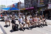 A group of people lined up sitting on chairs looking past the camera at a stage performance. Marrickville streetscape extends into the background including two temporary blue shade sails