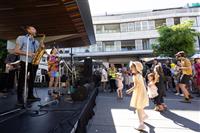 A side stage shot of a small band featuring a saxophonist, tambourine and electric guitar with a group of children and adults dancing to the music