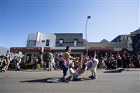 A wide streetscape featuring three physical theatre performers each wearing high vis orange vests and headbands, mid movement