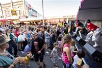 A side of stage shot with a group of three performers wearing white shoes and socks, variously coloured outfits and fringed masks, singing out to a large crowd gathered on a street