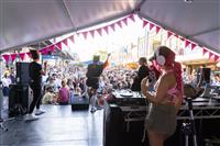 A back of stage shot with three performers, a DJ with bright pink braided hair and an auslan interpreter, playing to a large crowd of people gathered on a street