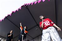 A low angle image looking up at a group of three performers in variously arranged sport-inspired outfits. The person singing raises their right arm