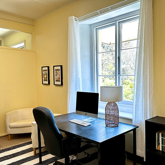 A desk and office chair with a computer faces out towards a window with a view of trees.