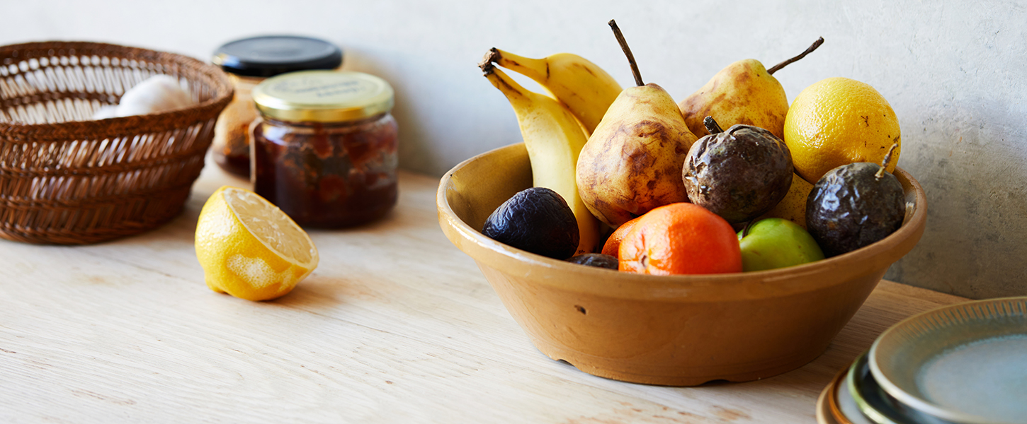 A bowl filled with overripe fruit next to an old half-lemon and two preserve jars