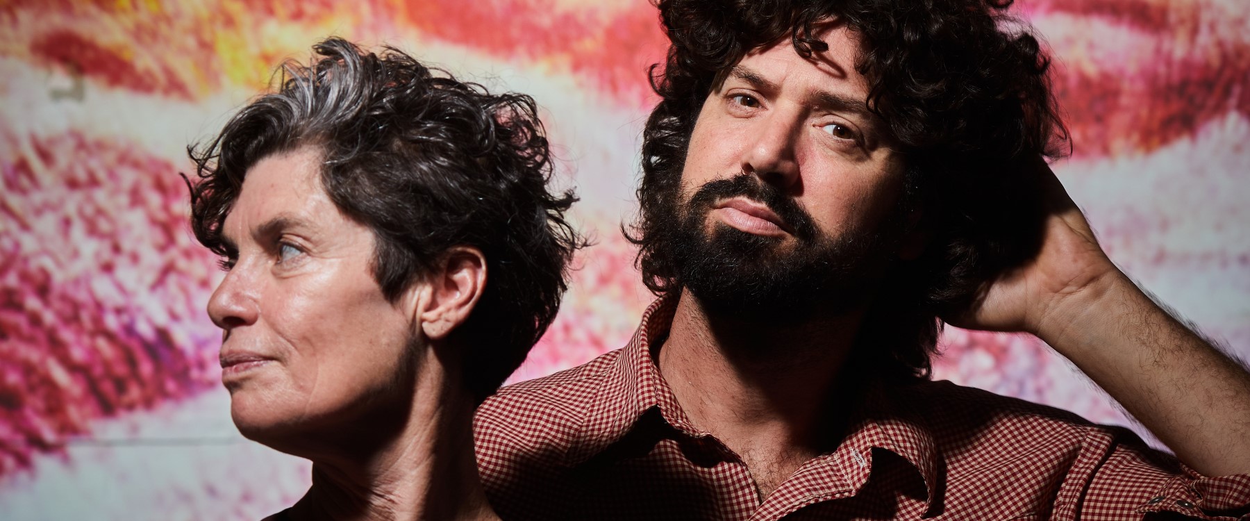 A close shot of two performers standing in front of a colourful digitally rendered image of reds, pinks, oranges and whites. The performer on the left has short dark and grey hair and looks off to the left. The performer on the right looks directly at the viewer and is wearing a red checkered shirt, large curly hair and dark facial hair