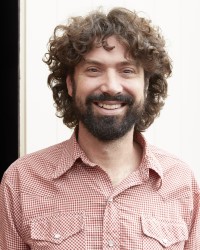 Headshot image of a person with a red checked shirt dark curly short hair and dark facial hair standing in front of a bright wall