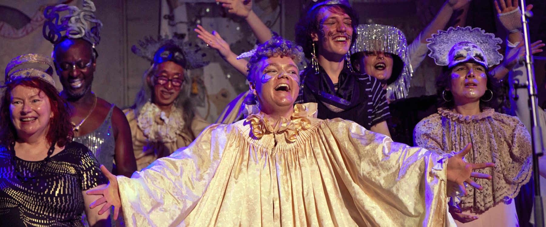 A group of eccentrically dressed choral performers face towards the viewer. They each wear various head dresses, collars, jewellery, costumes and makeup.