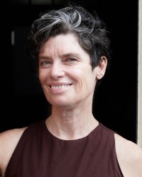 Headshot of a person with short dark and grey hair wearing a maroon singlet shirt standing in front of a darkened space