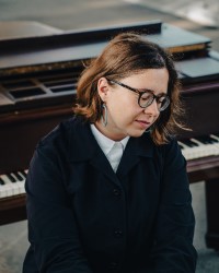 A composer sits in front of a piano wearing a white collared shirt and navy cardigan, looking down to the right. They wear circular tortoiseshell glasses and blue earrings with a brown bob