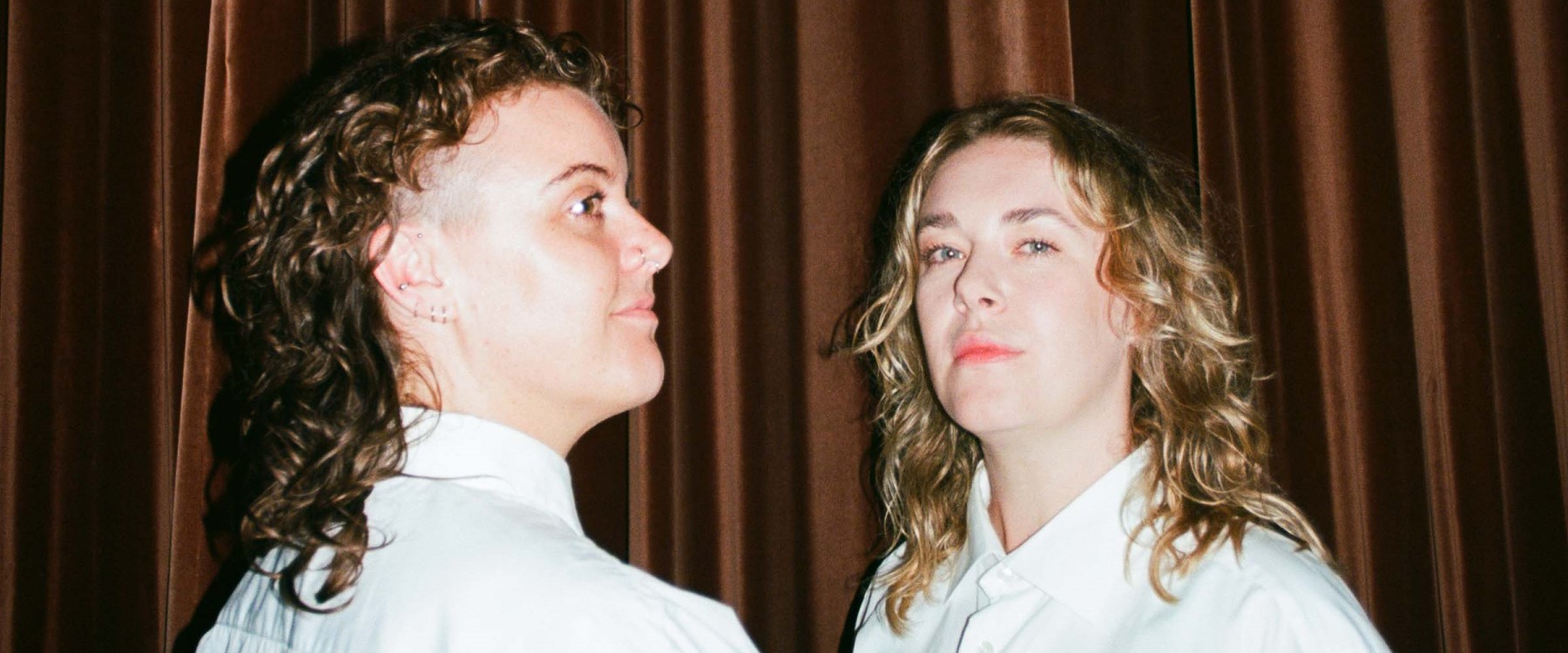 A close shot of two performers, both wearing white collared shirts against a brown velvet curtain backdrop. The left performer has brown wavy hair and various piercings, looks off to the right. The right performer looks at the viewer with blonde wavy hair.