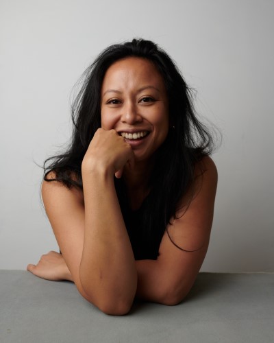 Headshot image of a person with dark hair and brown skin wearing a black t shirt, leaning towards the viewer over a desk with one hand coming up to their face to rest on their jaw