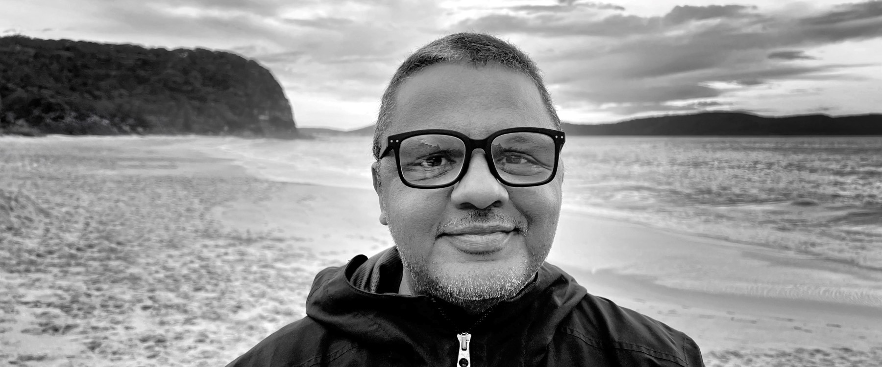 A black and white close shot of a person standing on a beach wearing a zipped up black jacket, black framed glasses, with short dark and grey hair and facial hair. In the background you can see waves rolling in and a bushland covered headland.