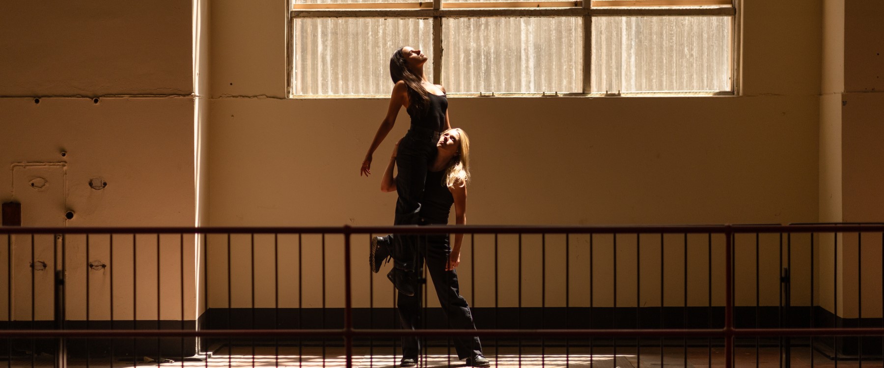 Two performers dressed in black, one lifting the other up towards a bright window, within a bright and warmly lit industrial space with a black balustrade in the foreground