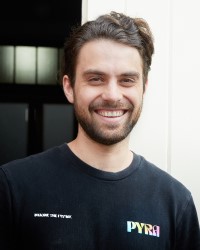 Headshot of a person with brown short hair and facial hair wearing a black t shirt with the word PYRA on the right standing in front of a bright wall and a darkened room
