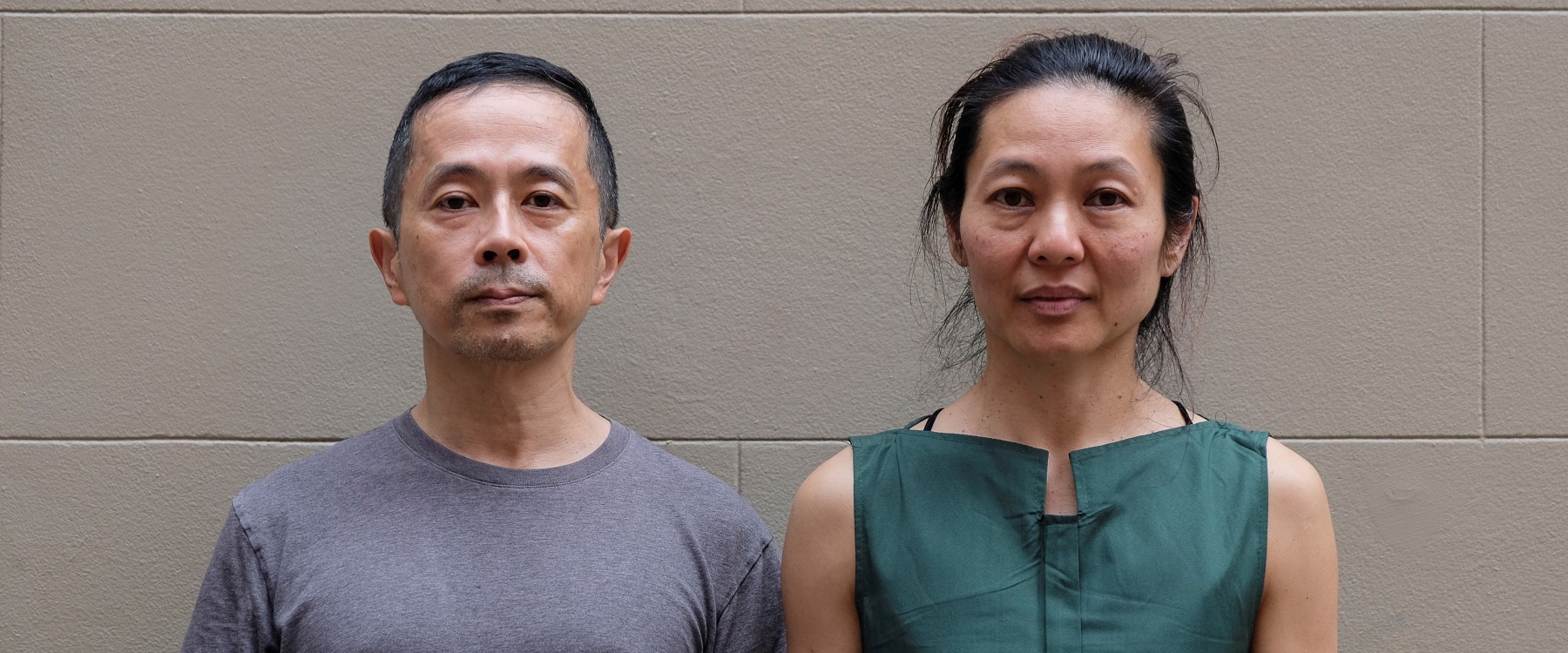 Two people stand against a grey wall looking straight at the camera with straight faces. One wears a grey t shirt with short black and grey hear, the other wears a green sleeveless shirt with tied back black hair