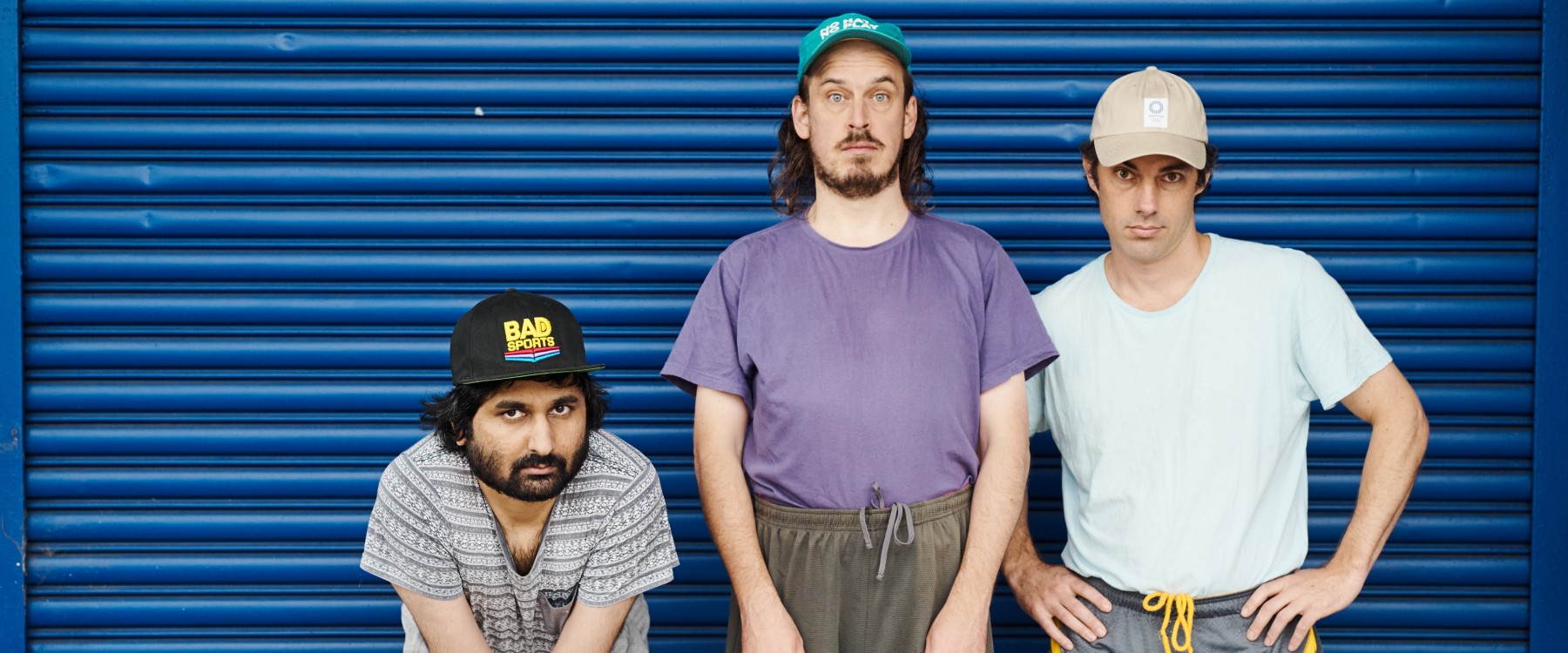 A group of three performers stand in front of a bright blue garage door. They each wear variously coloured and textured t shirts, drawstring shorts and caps. They all look directly and seriously at the viewer.