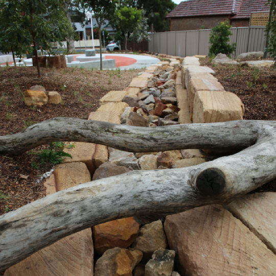 Sandstone blocks framing a line of jaggered multi-coloured rocks. A grey tree branch is laid over the top; green trees and a curved concrete structure is in the background