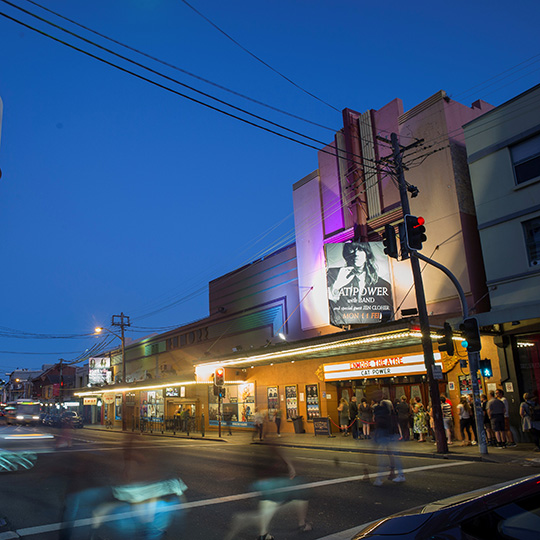 An art deco theatre at night with a line of people waiting outside.