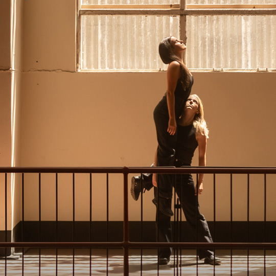 One person holds another person aloft at knee height in the stairway landing of an old building. Both people are looking upwards. The bright natural light from the opaque corrugated window behind the people adds to the drama of the scene.