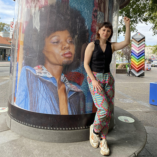 A woman with a raised left fist standing in front of an artwork encased in a clear cylinder.