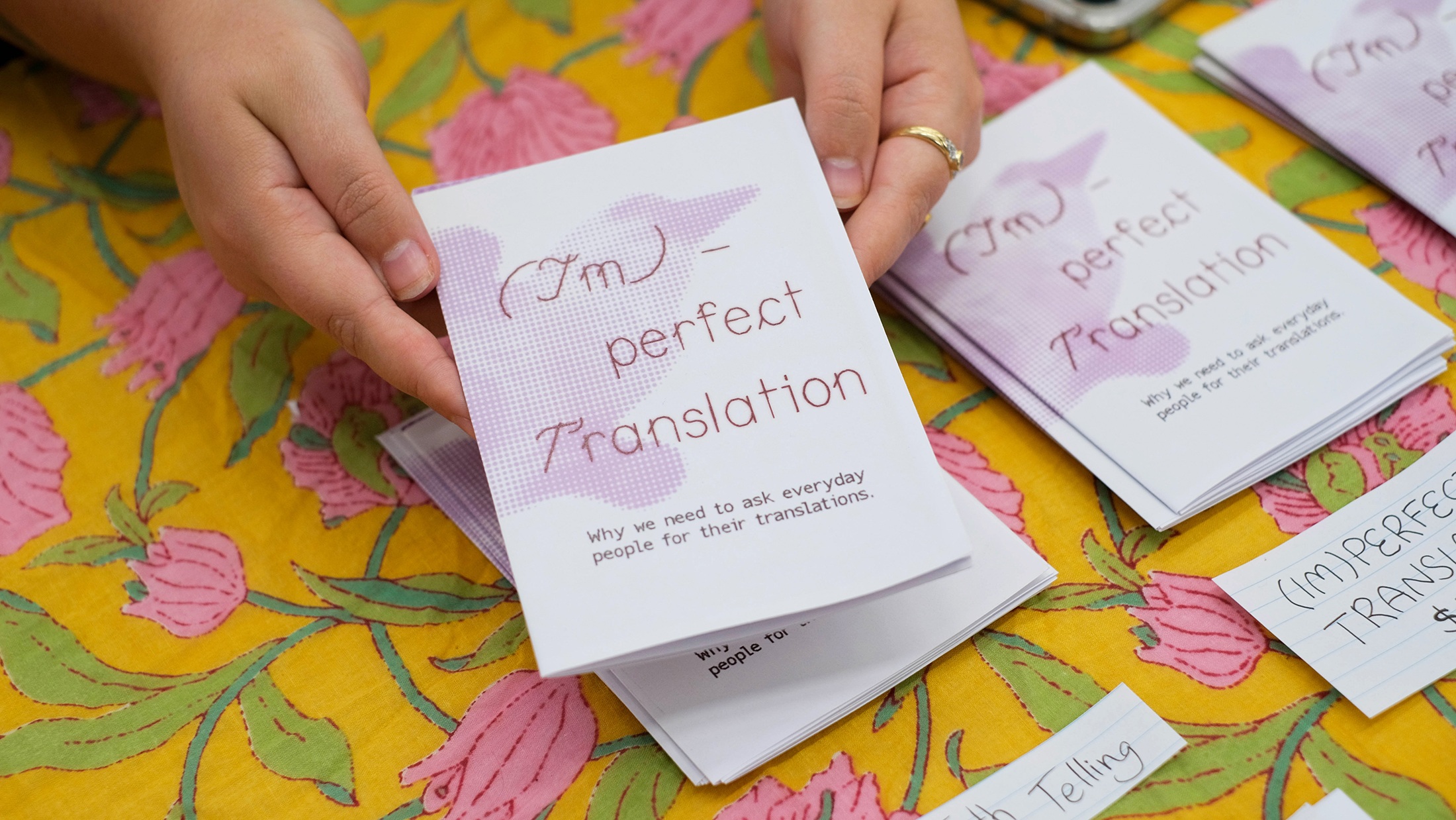 A person holding a folded pamphlet titled ‘(Im)perfect Translation’ with the subtitle ‘Why we need to ask everyday people for their translations.’ The pamphlet is placed on a bright yellow tablecloth with pink floral patterns, and several similar pamphlets and handwritten notes are scattered around
