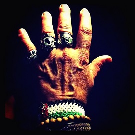 Photo of a left hand with many silver rings on pinky through to middle finger, the hand is outstretched, The hand has bracelets around the wrist, palestinian flag, wooden round beads and leather