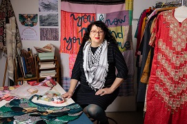 Photo of artist Nicole Barakat in her studio, she sits on a stoll and wears a Keffiyeh, she is surrounded by art making materials and is sitting beside a table that has sewing materials like pin cushions and intricate mermaid like material in a fish scale design with rounded pieces of mixed material in blues, greens and gold threads overlay each other