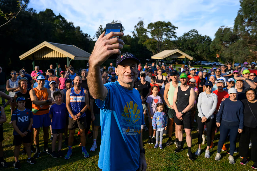 A man holding his camera up in 'selfie' position to photograph himself and a large group of other people in an urban park.