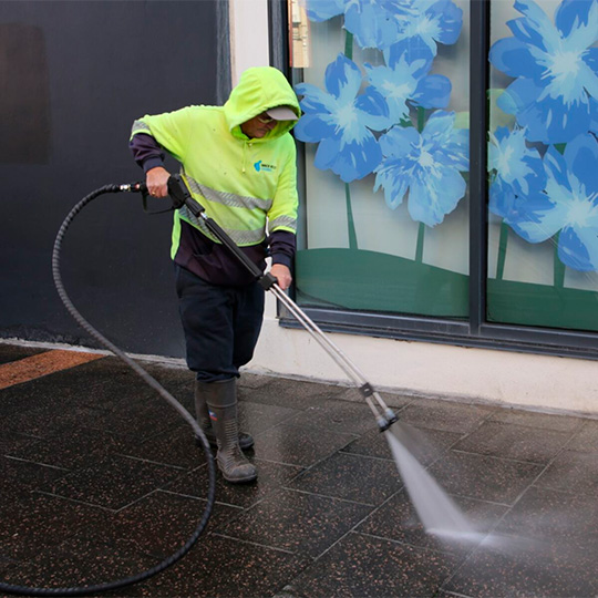 A worker in boots and high-visibility gear cleans a main street stone-tile footpath with a high-pressure water hose.