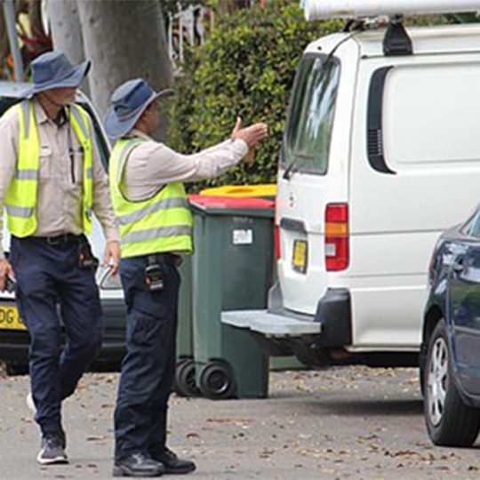 Two rangers in high-visibility vests investigate a van parked across a footpath.