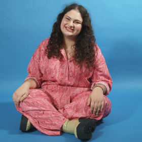 Michelle wears pink jumpsuit, smiling. light blue backdrop