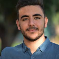 Young man with tanned skin, dark features and dark hair wearing a blue button up shirt in a natural setting