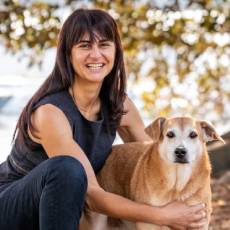 Woman with tanned skin and long black hair wearing a black singlet and jeans crouching in a natural setting hugging a blonde coloured dog