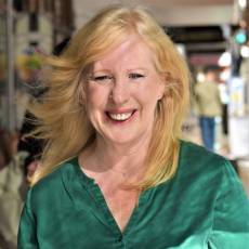 Woman with fair skin and long blonde hair wearing an emerald green blouse in front of a street scape