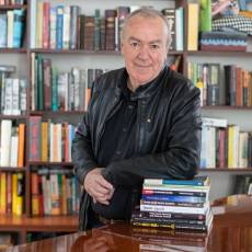 Man with fair skin and short grey hair wearing a black button up shirt and black leather jacket leaning on a pile of books placed on a polished timber table in front of a large bookcase filled with books