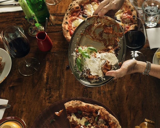 A pair of hands holding a shared pasta dish above a pizzeria table setting.