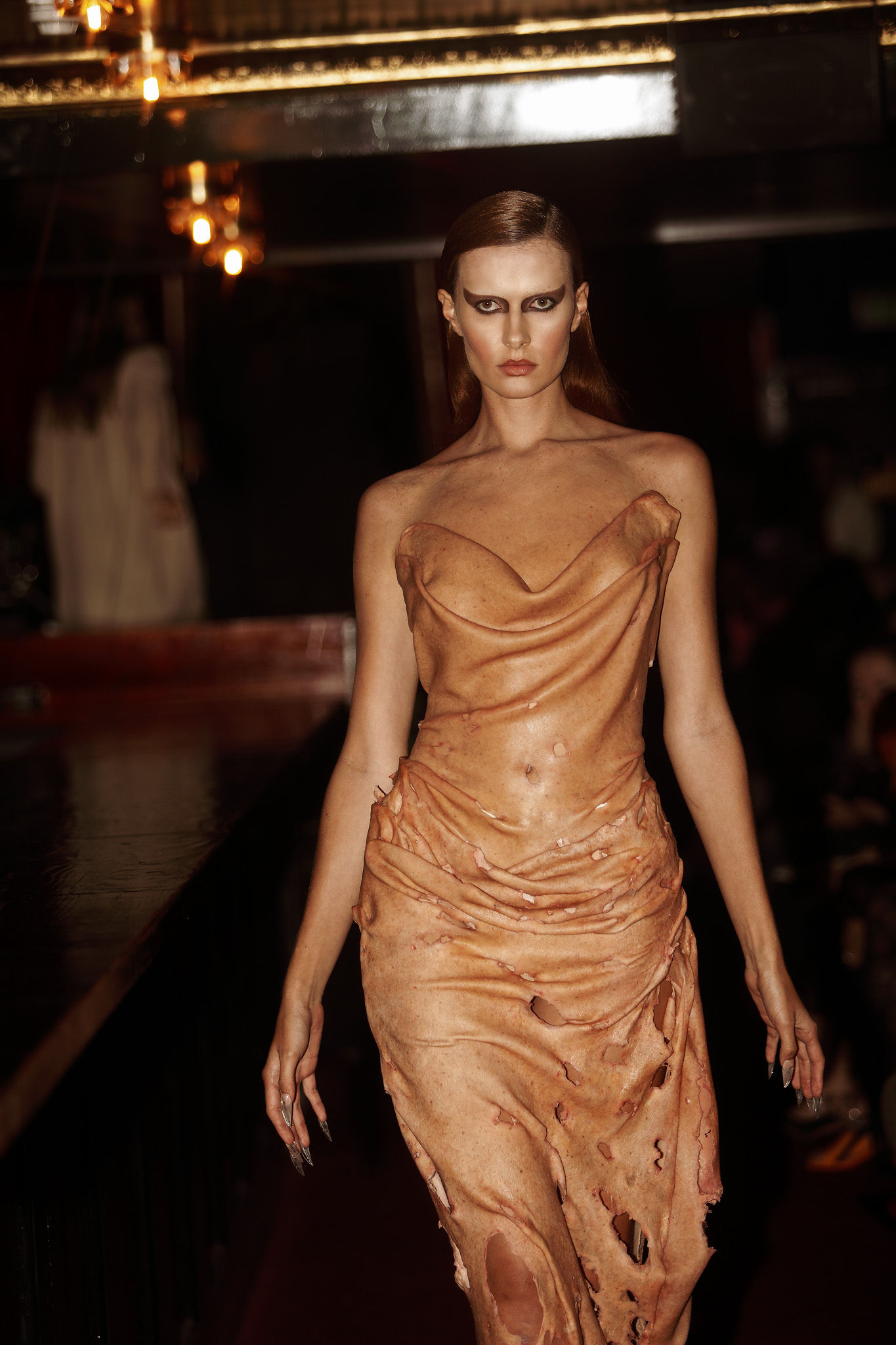 Janet Anderson walks a red carpet runway in a bronze dress with holes. 