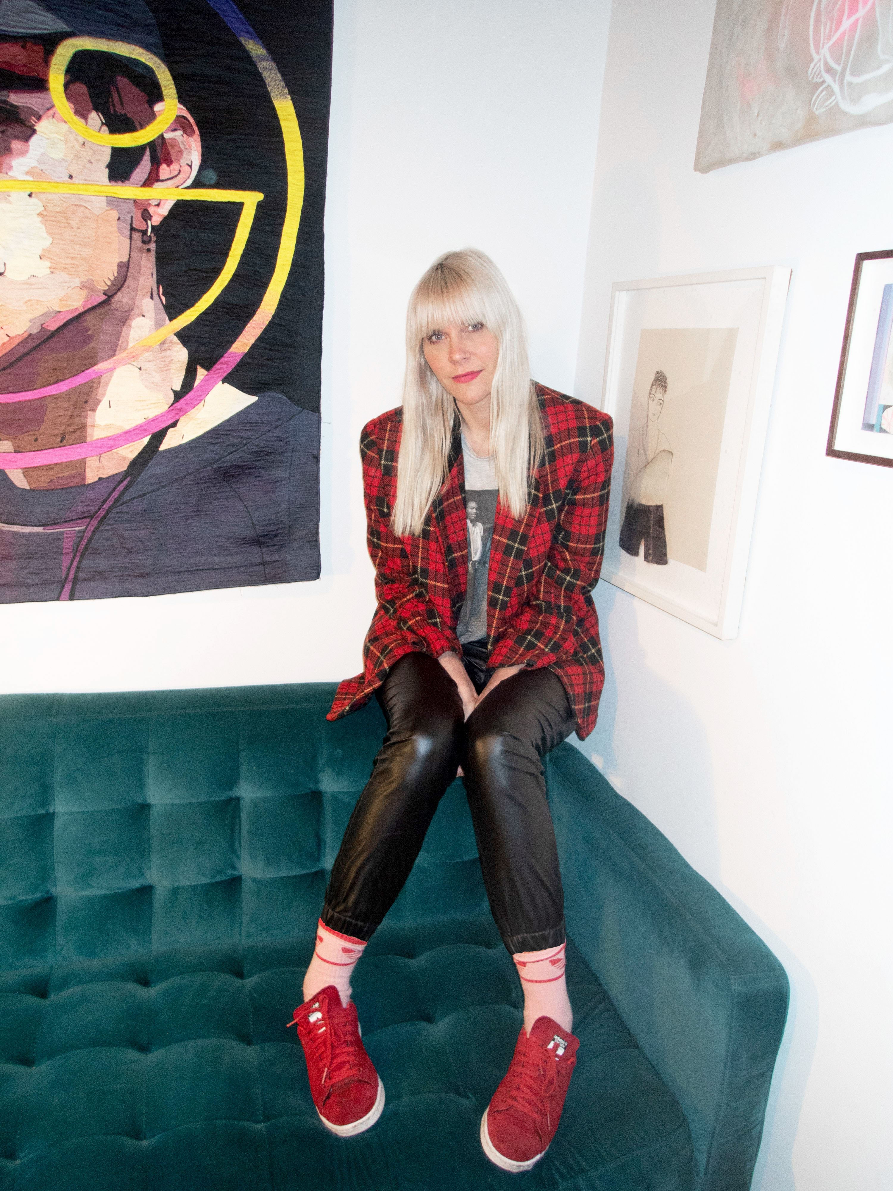 Sam Watson-Wood sits on the corner of a couch surrounded by artworks and looks into the camera. 
