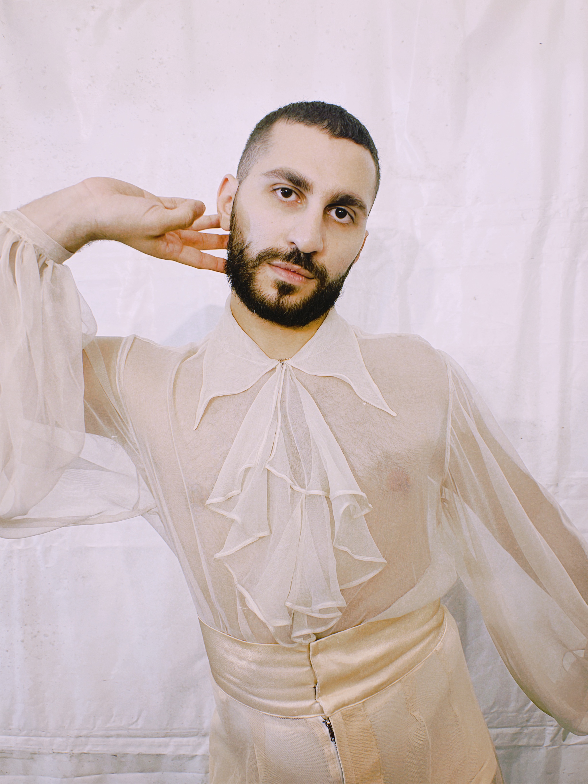 Xander Khoury posted in a sheer white top in front of a white background.