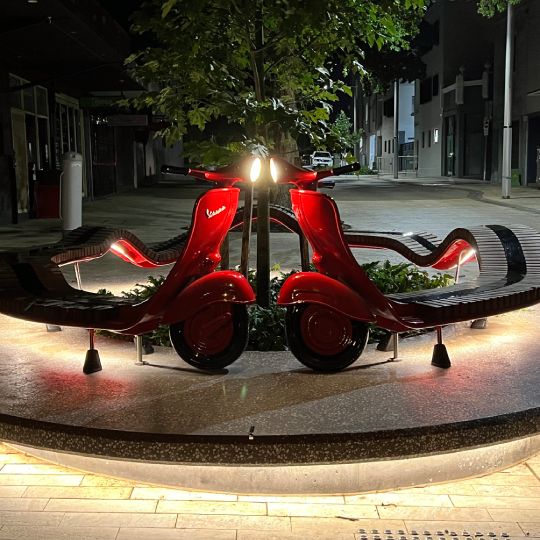 A sculpture at night with two red scooters turned into a bench with lights underneath surround a tree. 